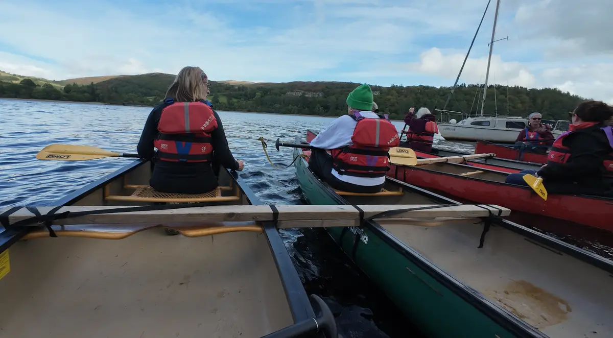 Women in red buoyancy aids paddling rafted-up canoes across the lake